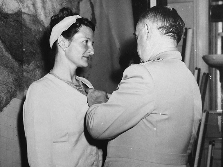 black and white photos of a dark haired woman receiving medal of honour from a man with his back turned to the camera