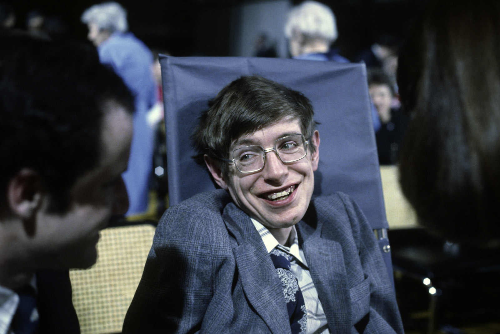 Stephen Hawking dressed in blue suit with matching blue tie smiling into camera