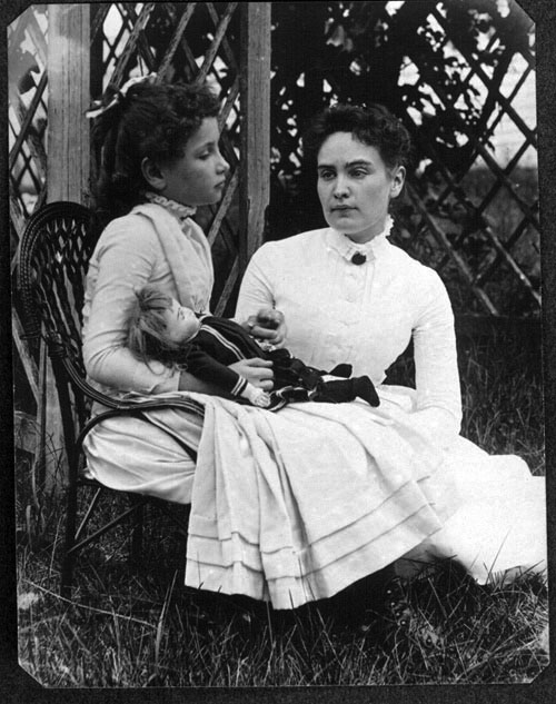 black and white picture of young girl sitting in wicker chair with older woman looking on. Both wearing white dresses.