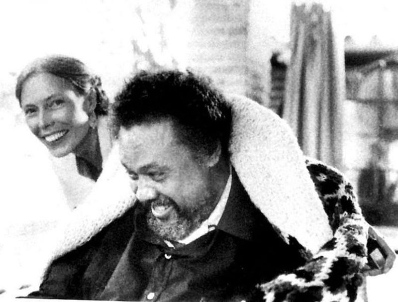 a smiling Joni Mitchell with her arms around a sitting Charles Mingus