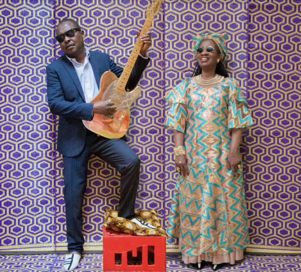 African man in blue suit holding gutar accompanied by African woman wearing colourul floral pattern dress