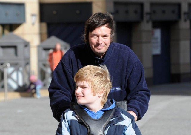 Ian Ranking wearing dark jacket pushing his son Kit who is sitting in a wheelchair. Kit has light brown hair, is wearing a blue jacket and looking to his right.