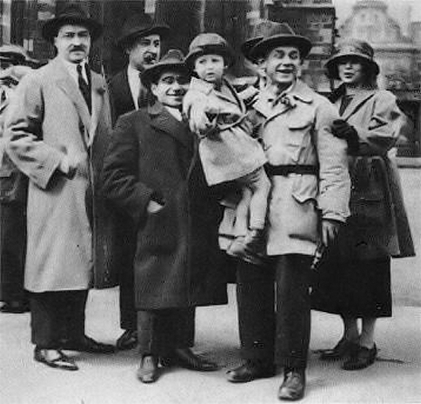 black & white photo of 6 adults. One of the adults is holding a child in his arms. On his left is a smiling man of short stature wearing a dark overcoat.
