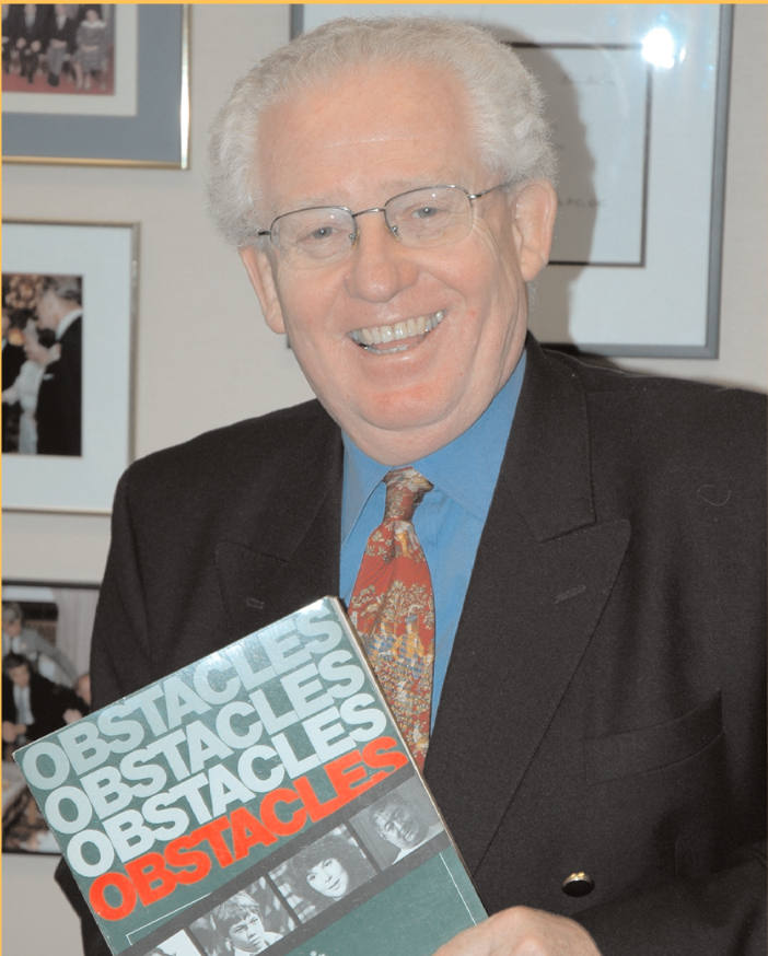 smiling man with grey hair, wearing glasses with a blue shirt and red pailsley tie. He is holda copy of the Obstacles Report