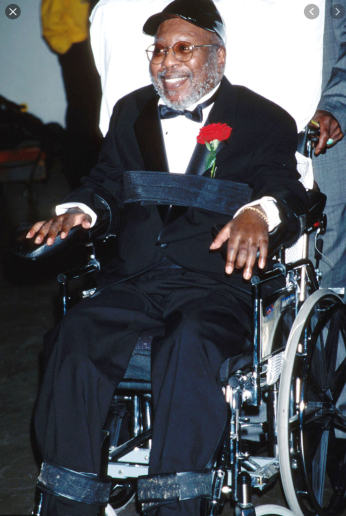 Photo of Curtis Mayfield sitting in his wheelchair. He is dressed in a tuxedo with a red carnation in his lapel.