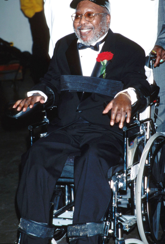 Photo of Curtis Mayfield sitting in his wheelchair. He is dressed in a tuxedo with a red carnation in his lapel.