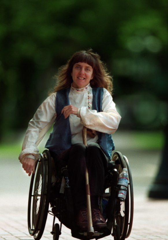 Susan Sygall rolling in her wheelchair. She is wearing a white blouse with a blue scarf around her neck.