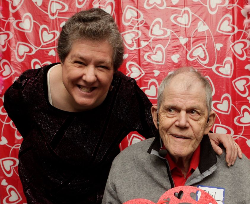Photo of Phill Allen seated and smiling. His wife Wendyis standing beside him with her hand on his shoulder. Red and white hearts are in the background.